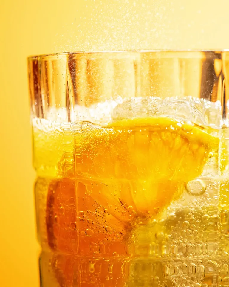 Produktfotografie eines Softdrinks mit Orangenscheibe und Eiswürfeln im Detail vor orangfarbigen Hintergrund. Klaus Ohlenschläger, Studio für Produktfotografie